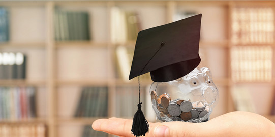 Scholarship A transparent piggy bank full of coins and with a doctoral cap is held in front of a book shelf.